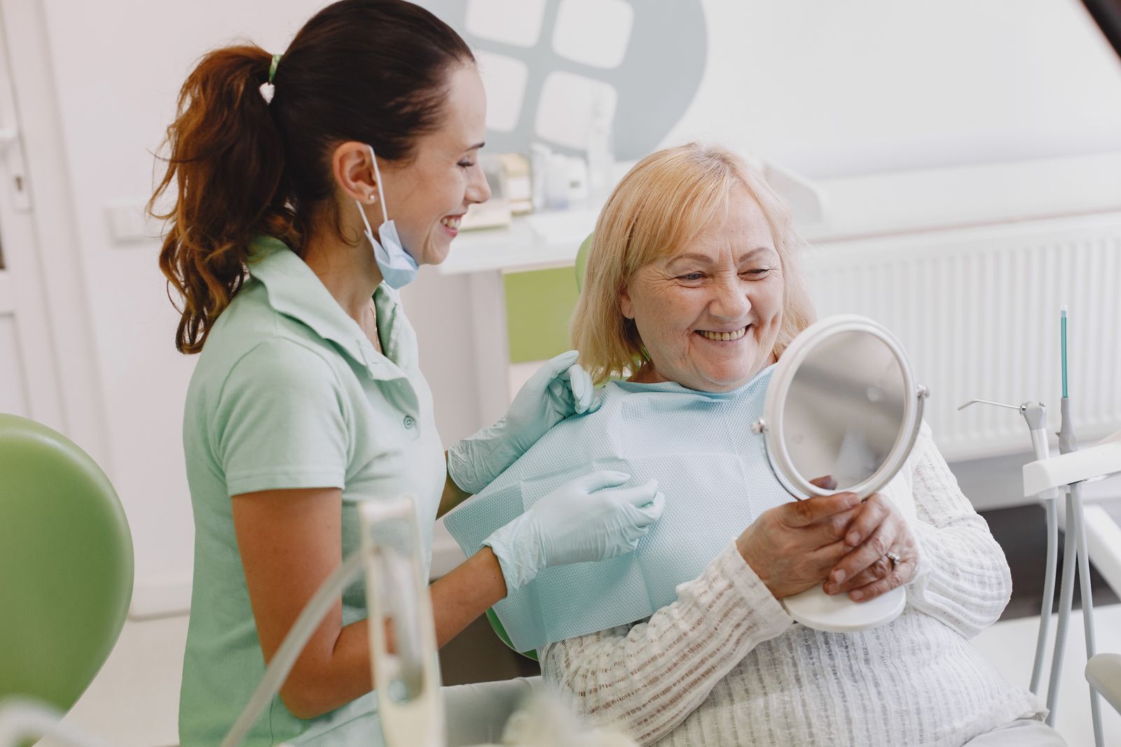 A patient smiling at herself in a hand mirror beside a hygienist